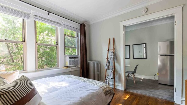 A bedroom with a bed, a radiator, a window with curtains, and a view of the outdoors. - Rustic Studio Apartment in the Heart of Lincoln Square (Chicago)