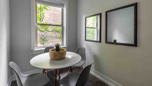 A room with a round table, four chairs, a wooden planter with green plants, and two windows with blinds. - Rustic Studio Apartment in the Heart of Lincoln Square (Chicago)