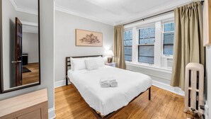 A hotel room with a large bed, a bedside table with a lamp, a window with curtains, and a radiator. - Beautiful and Comfy 1BR in Lincoln Square (Chicago)
