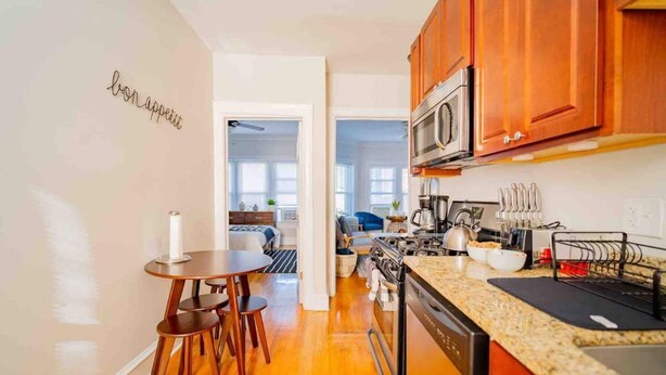 A modern kitchen with wooden cabinets, a built-in microwave, and a granite countertop. There is a dining area with a round table and chairs. - Stylish and Comfy Retreat 10 Minutes from Wrigley (Chicago)