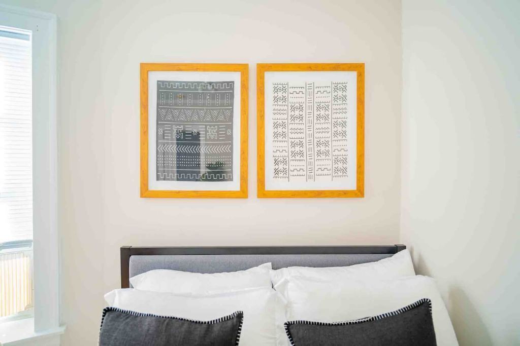 A bedroom with a bed, two framed artworks on the wall, and a window to the left.