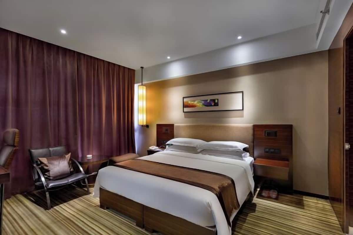 Quarto de hotel com cama grande, uma cadeira, mesa de cabeceira e uma obra de arte na parede.