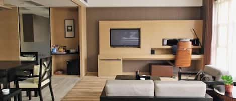 A modern hotel room with a sofa, a dining table, chairs, a TV, and a small plant.