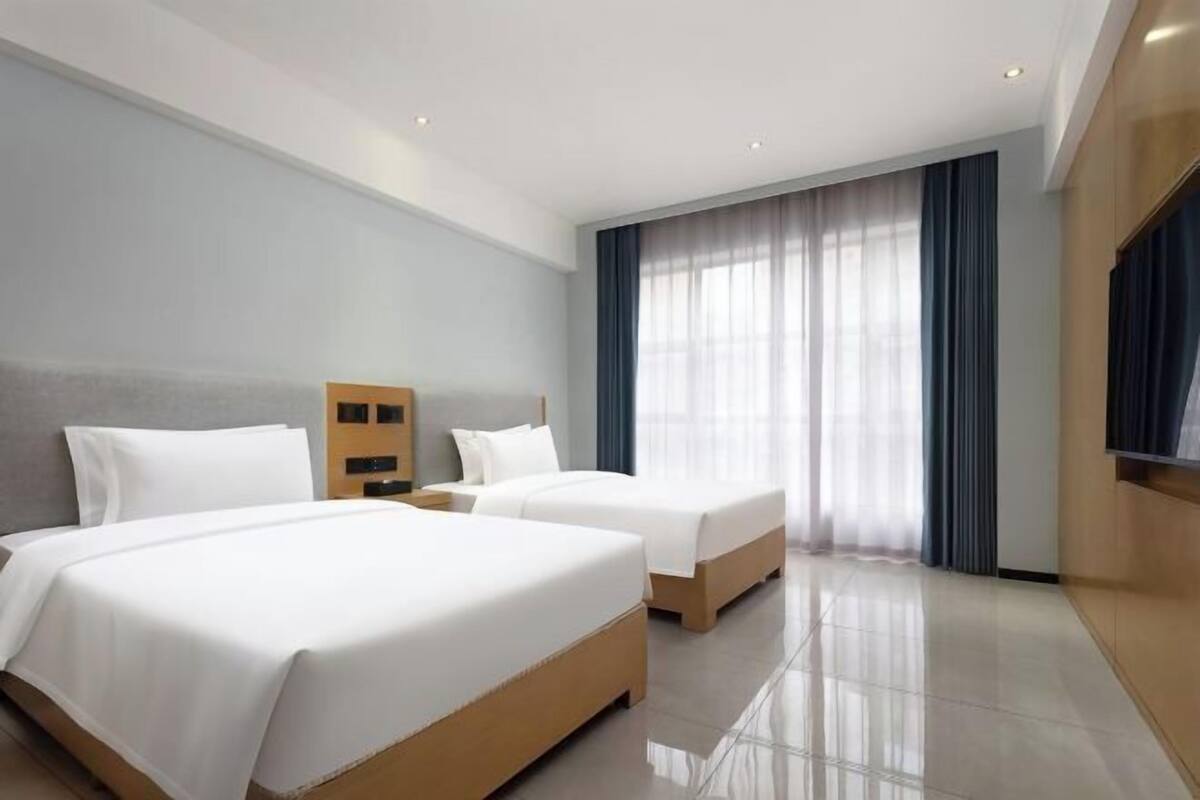 A hotel room with two beds, a wooden headboard, a nightstand, a TV, and a large window with curtains.