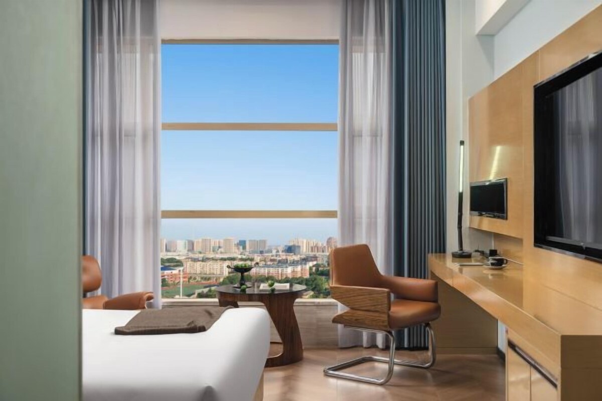 Camera d'albergo moderna con una grande finestra che si affaccia sulla città, una TV a schermo piatto montata a parete e un'area salotto con tavolo e sedie.