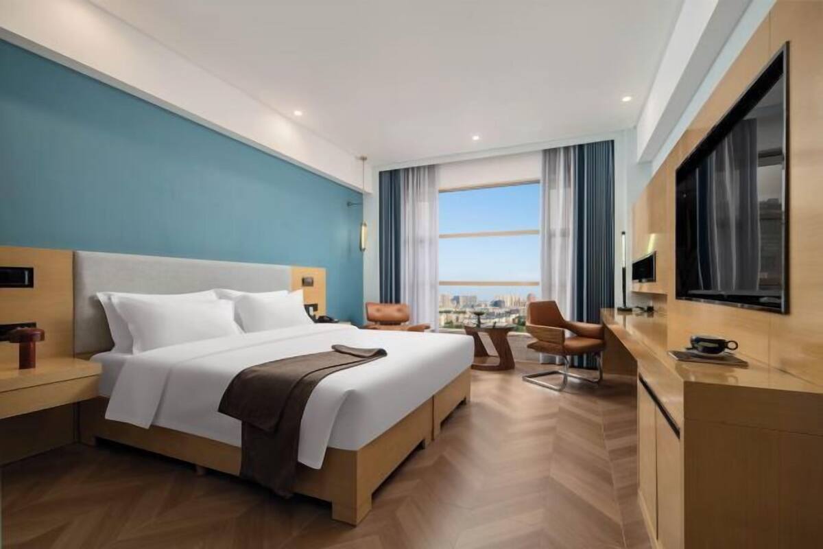 A modern hotel room with a large bed, a desk, a chair, and a view of the city.