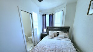 A bedroom with a bed, a nightstand, a window with curtains, and a door leading to a bathroom.