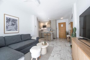Una sala de estar moderna con un sofá gris, una mesita redonda pequeña y un taburete blanco y mullido. Se ve una cocina con barra al fondo.