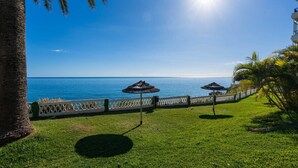 A grassy area with a white fence, two thatched umbrellas, and a view of the sea. - Acapulco Playa 302 by Casasol (Nerja)