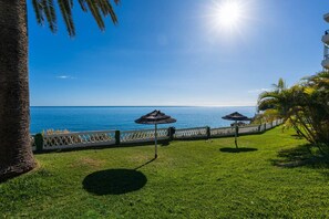 Un área con césped, una cerca blanca, dos sombrillas de paja y vista al mar.