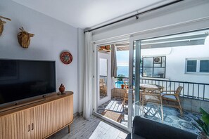 Un balcón con vistas al mar, un televisor sobre un mueble de madera y mobiliario de mimbre.