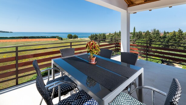 Uma área de refeições ao ar livre coberta, com mesa e cadeiras, com vista para o mar e vegetação.