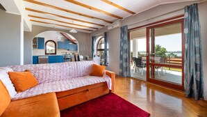 Uma sala de estar moderna com sofá, tapete vermelho, vigas de madeira, cozinha ao fundo e grandes portas de vidro deslizantes para área externa.