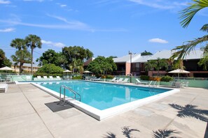 A rectangular swimming pool with a metal ladder, surrounded by lounge chairs and umbrellas, with palm trees and a building in the background. - Mariners House @ Landings of Largo (Key Largo)