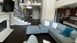 A spacious living room with a sectional sofa, a flat-screen TV, a fireplace, and a staircase leading to an upper level. - Newly Listed Oasis (Pearland)