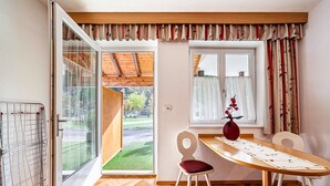 Ein Zimmer mit Holzboden, einem Esstisch mit Stühlen, einem Fenster mit Vorhängen und Blick auf eine grüne Außenanlage.