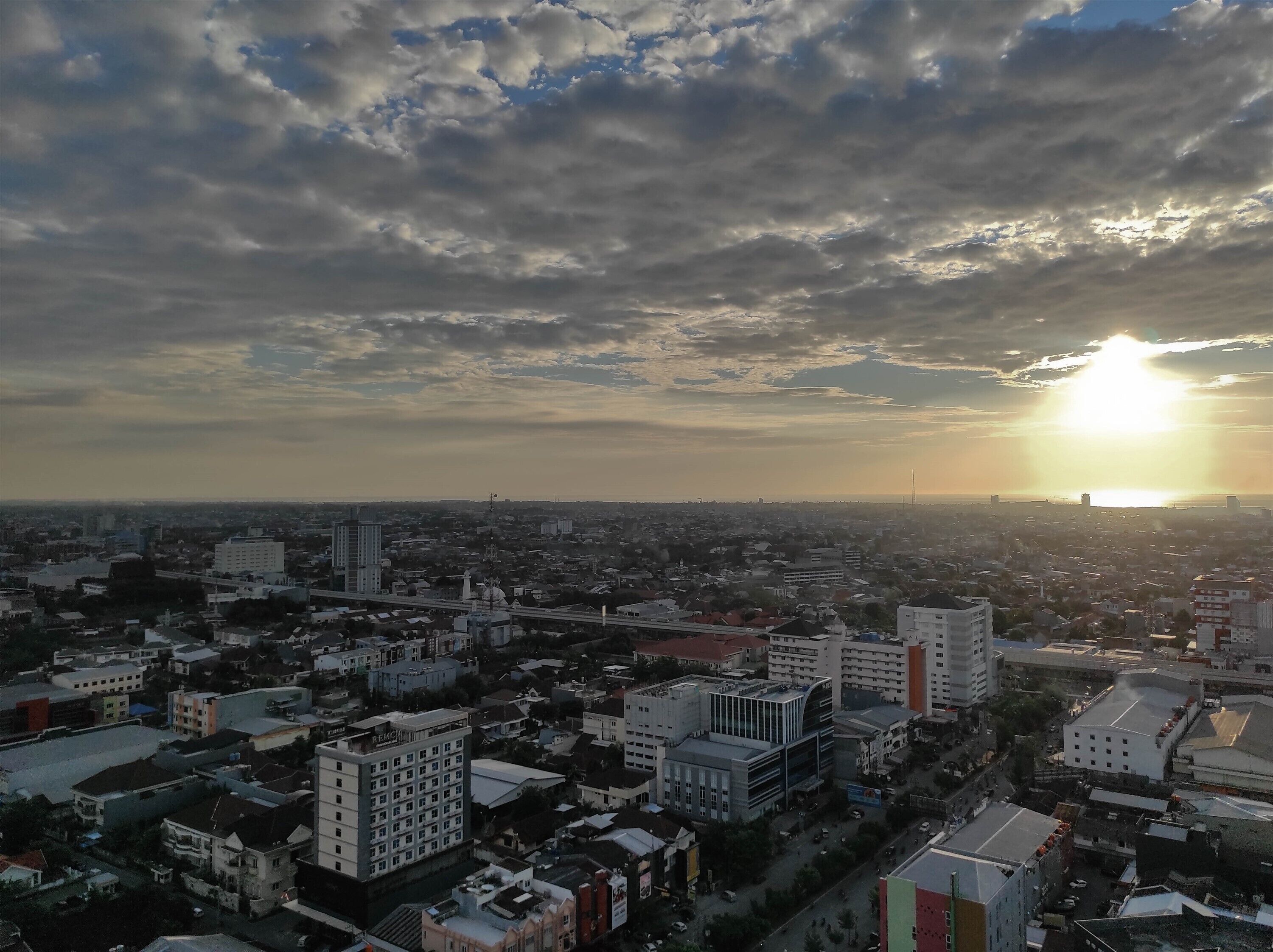 A cityscape with a variety of buildings, including high-rise and low-rise structures, under a partly cloudy sky with the sun setting or rising.