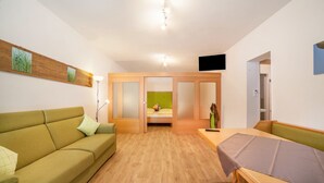 Una habitación con un sofá verde, una cama de madera, una mesita pequeña y un televisor montado en la pared.