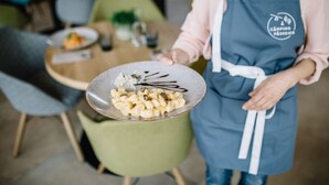Una persona sosteniendo un plato con comida, usando un delantal azul con un logotipo, en un área de comedor con sillas y una mesa.