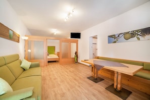 A room with a green sofa, a wooden dining table, a TV mounted on the wall, and a wooden floor.