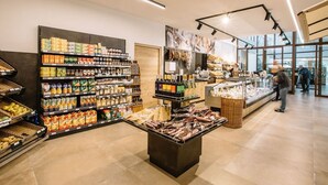 Interior de una tienda de comestibles con estantes llenos de diversos productos, una vitrina central y vista al exterior a través de grandes ventanales.