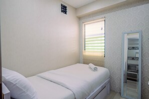 A small, neatly made bed with a white mattress and pillows, a window with blinds, and a mirror with a decorative frame. - Simple And Cozy Living 2Br At Cibubur Village Apartment (Depok)