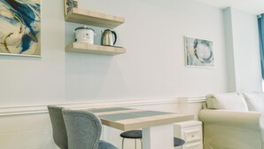 Een moderne woonkamer met een bank, een eettafel en een stoel. Twee houten planken met keukengerei hangen aan de muur.