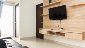 A modern living room with a flat-screen TV mounted on a wooden wall, a black leather sofa, and a sliding glass door leading to a balcony. - Comfortable 2Br Apartment At Mustika Golf Residence (Cikarang)