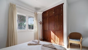 Une chambre avec une grande armoire, un fauteuil en osier et un lit agrémenté d’oreillers à rayures.