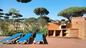 Une terrasse avec deux chaises longues, un barbecue en briques et un grand arbre.