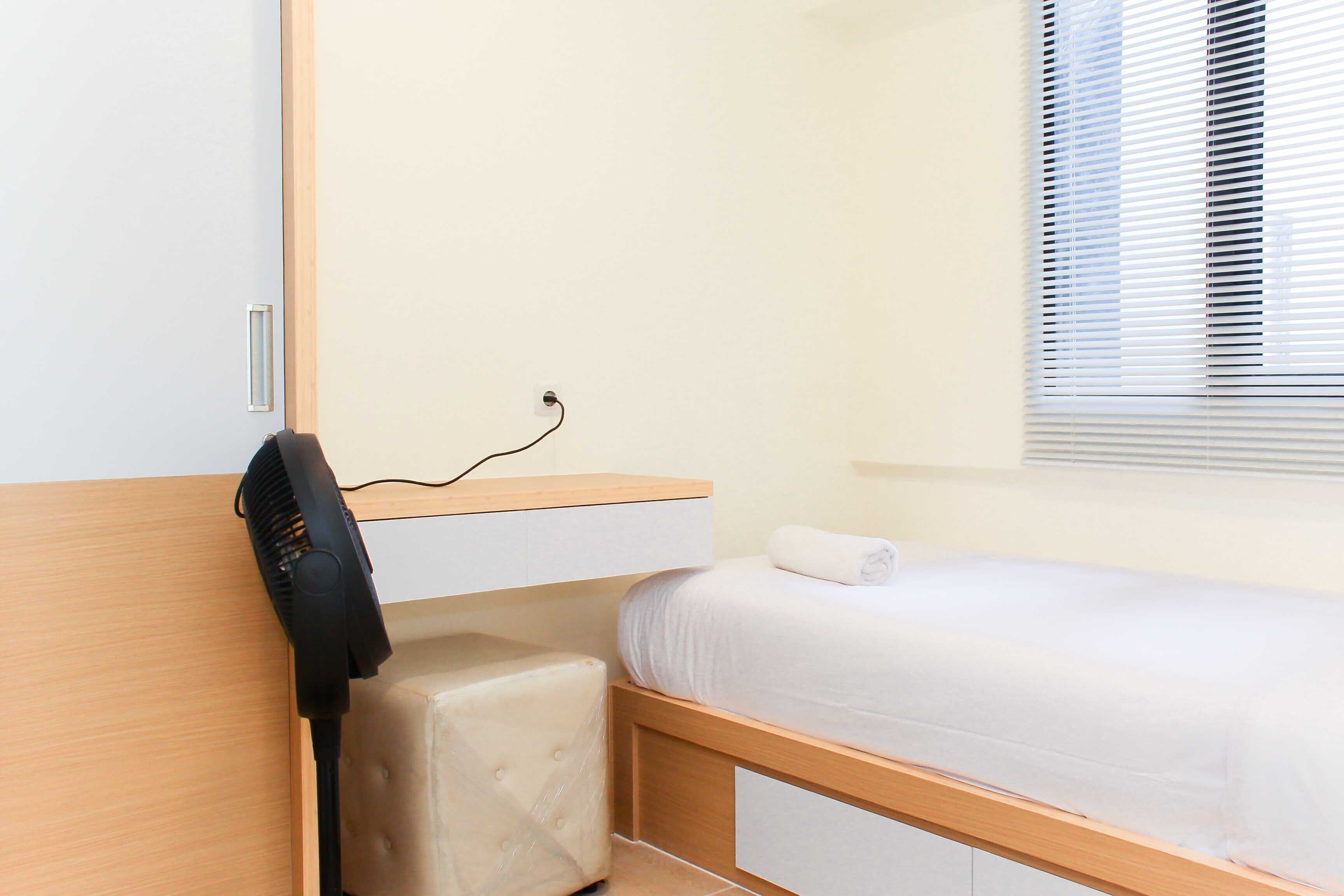 Un dormitorio pequeño y ordenado con una cama individual, una mesita de noche, una silla y un ventilador de pared.