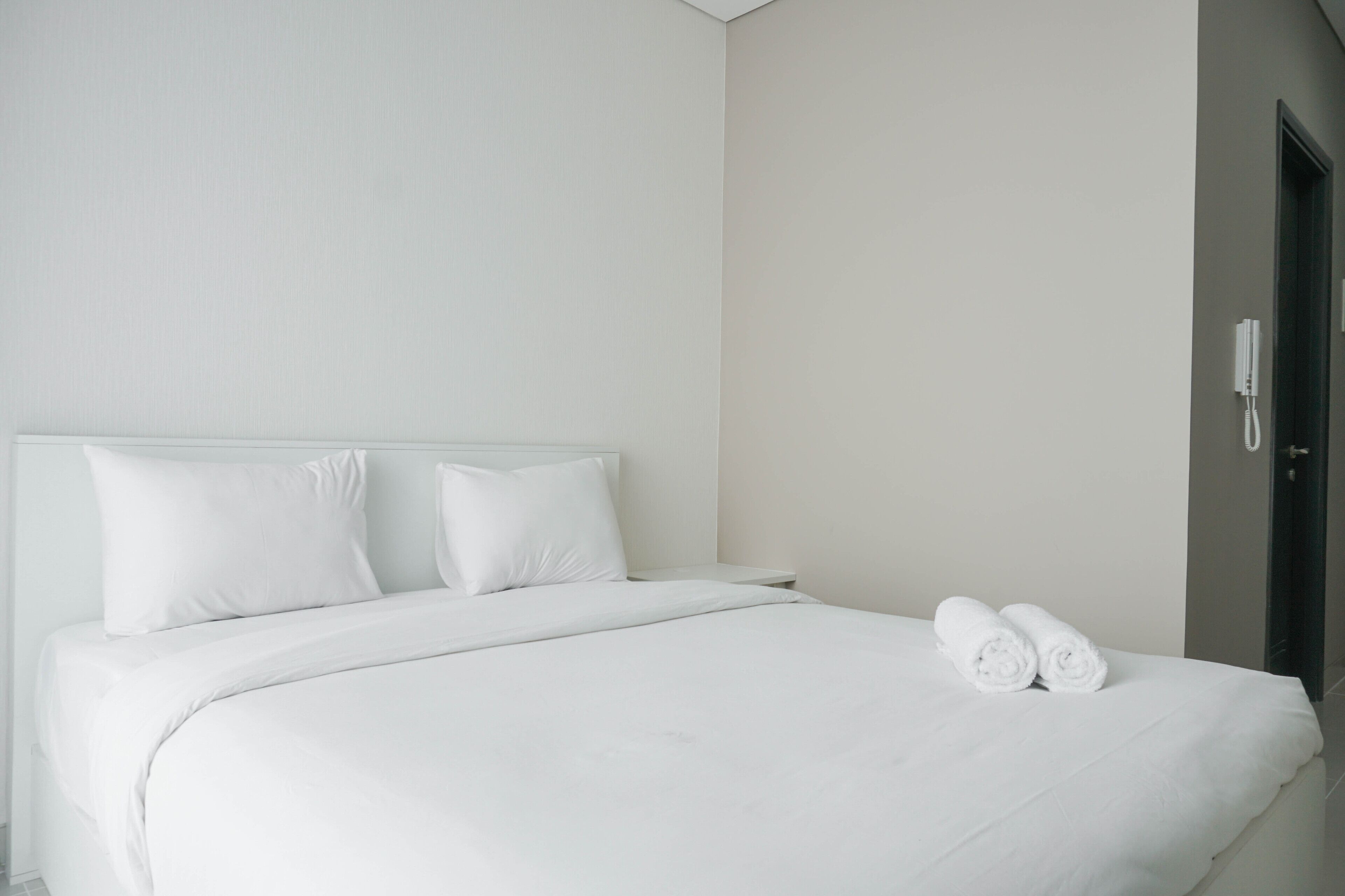 A neatly made bed with white linens and pillows, a pair of rolled towels on the bed, and a white telephone on the wall.