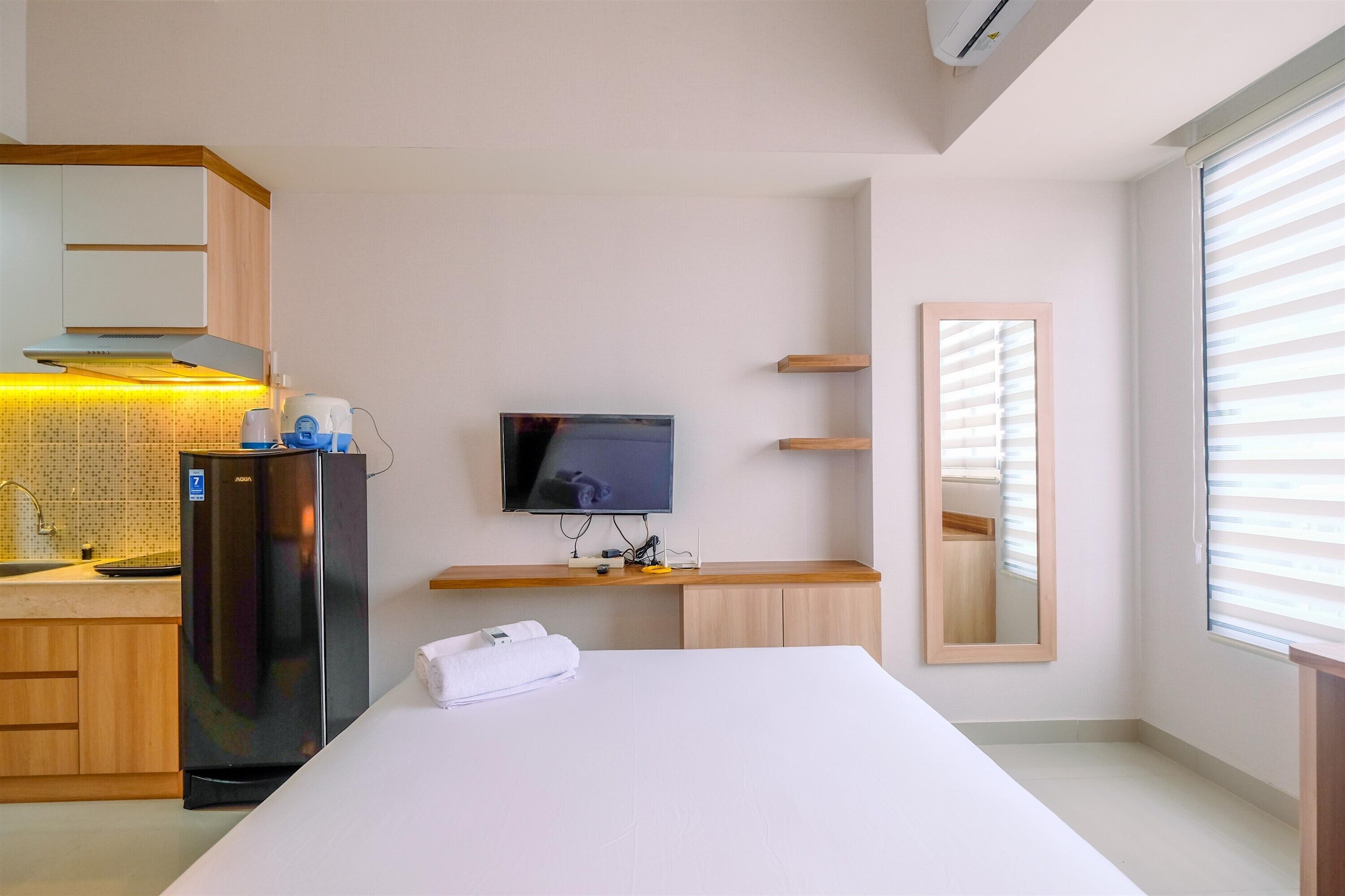 A hotel room with a bed, a TV, a refrigerator, a kitchen area, and a window with blinds.