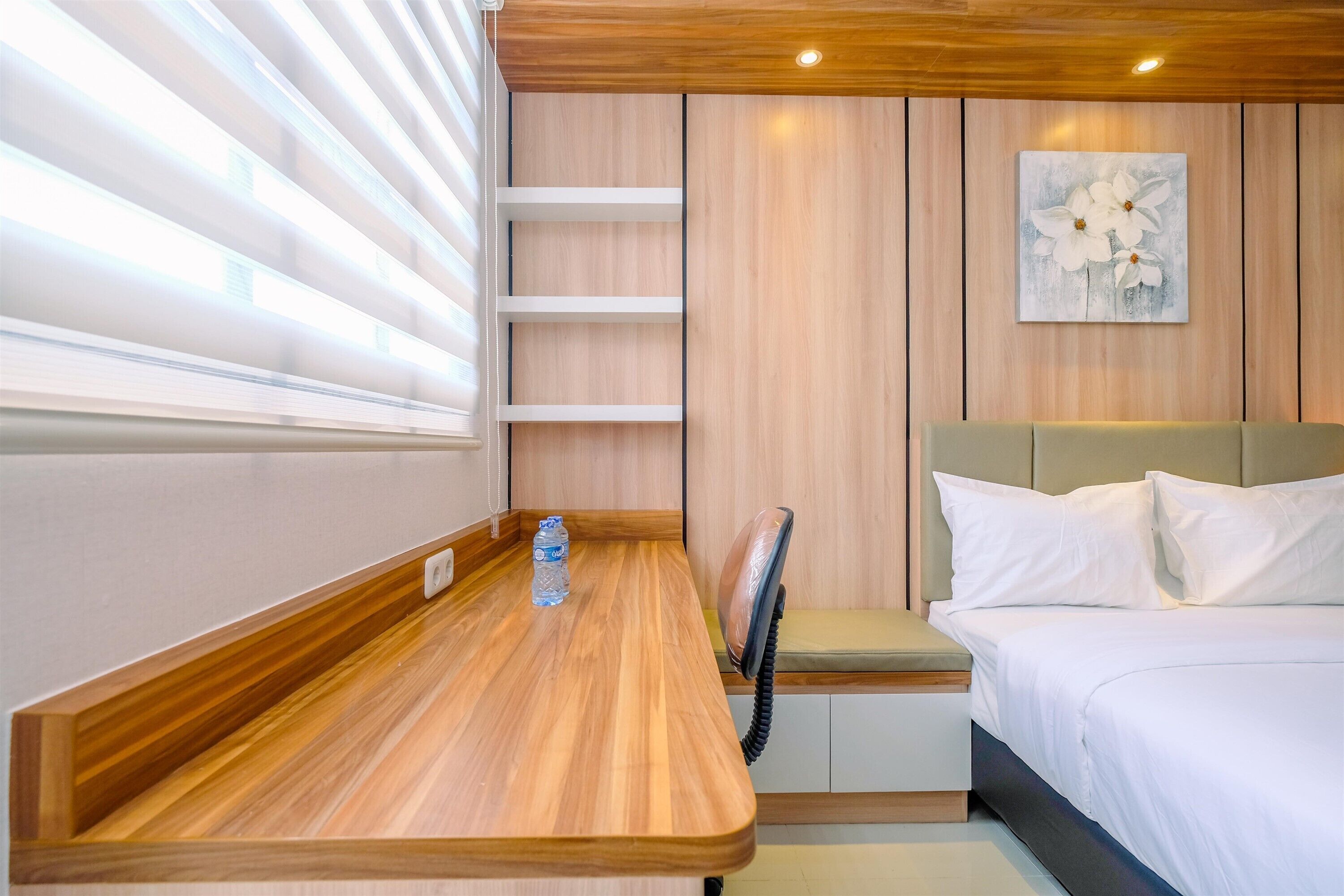 A modern hotel room with a wooden desk, a bed with white linens, a painting on the wall, and a window with blinds.