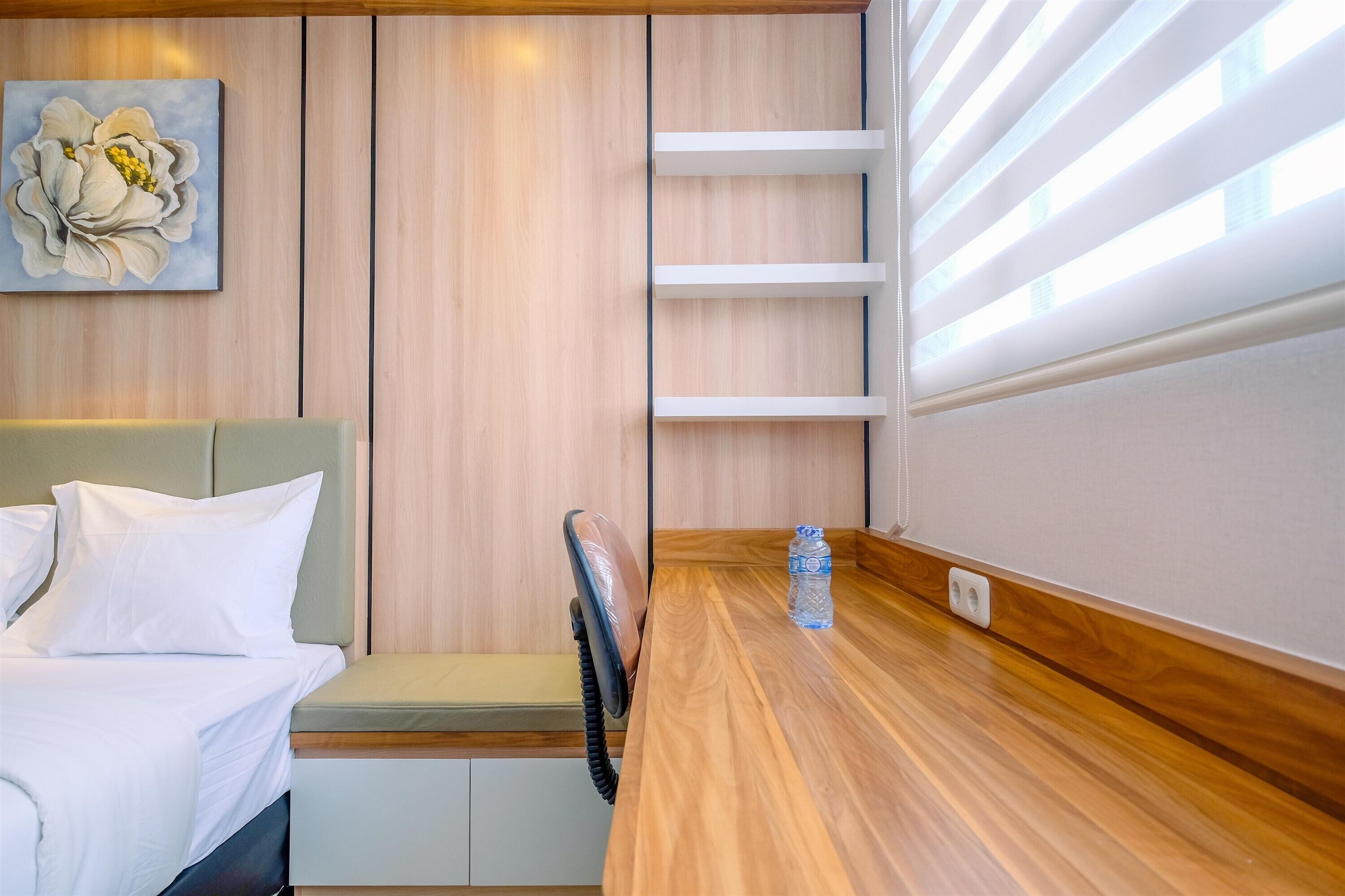 Une chambre moderne avec une tête de lit en bois, un lit recouvert de linge de lit blanc, un banc et une table en bois sur laquelle se trouve une bouteille d’eau.