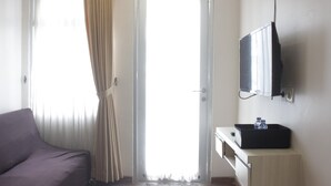 A hotel room with a bed, a television, a small table with a water bottle, and a door leading to a bright area. - Pool View 2Br Apartment At The Jarrdin Cihampelas (Bandung)