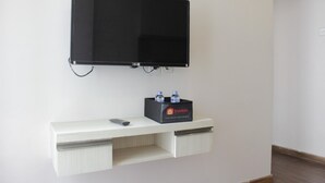 A flat-screen TV mounted on the wall above a white shelf with a remote control and a box labeled "travelio." - Pool View 2Br Apartment At The Jarrdin Cihampelas (Bandung)