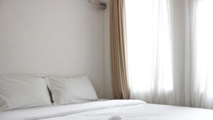 A neatly made bed with white linens and a single pillow, a white bedside table with a small object on it, and sheer curtains allowing natural light into the room. - Pool View 2Br Apartment At The Jarrdin Cihampelas (Bandung)
