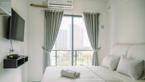 A hotel room with a bed, a TV mounted on the wall, and a window with curtains. - Comfortable Studio With Pool View At Sky House Bsd Apartment (Pagedangan)