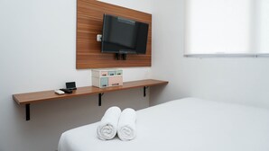 A hotel room with a bed, a TV mounted on the wall, a wooden shelf with a remote control and a box, and a window with blinds.