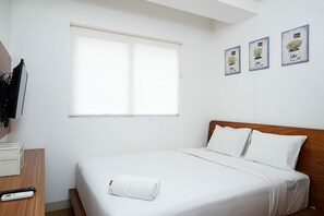A neatly made bed with white linens, a wooden headboard, and a white towel on the foot of the bed. There is a window with blinds, a television mounted on the wall, and three framed pictures on the wall.