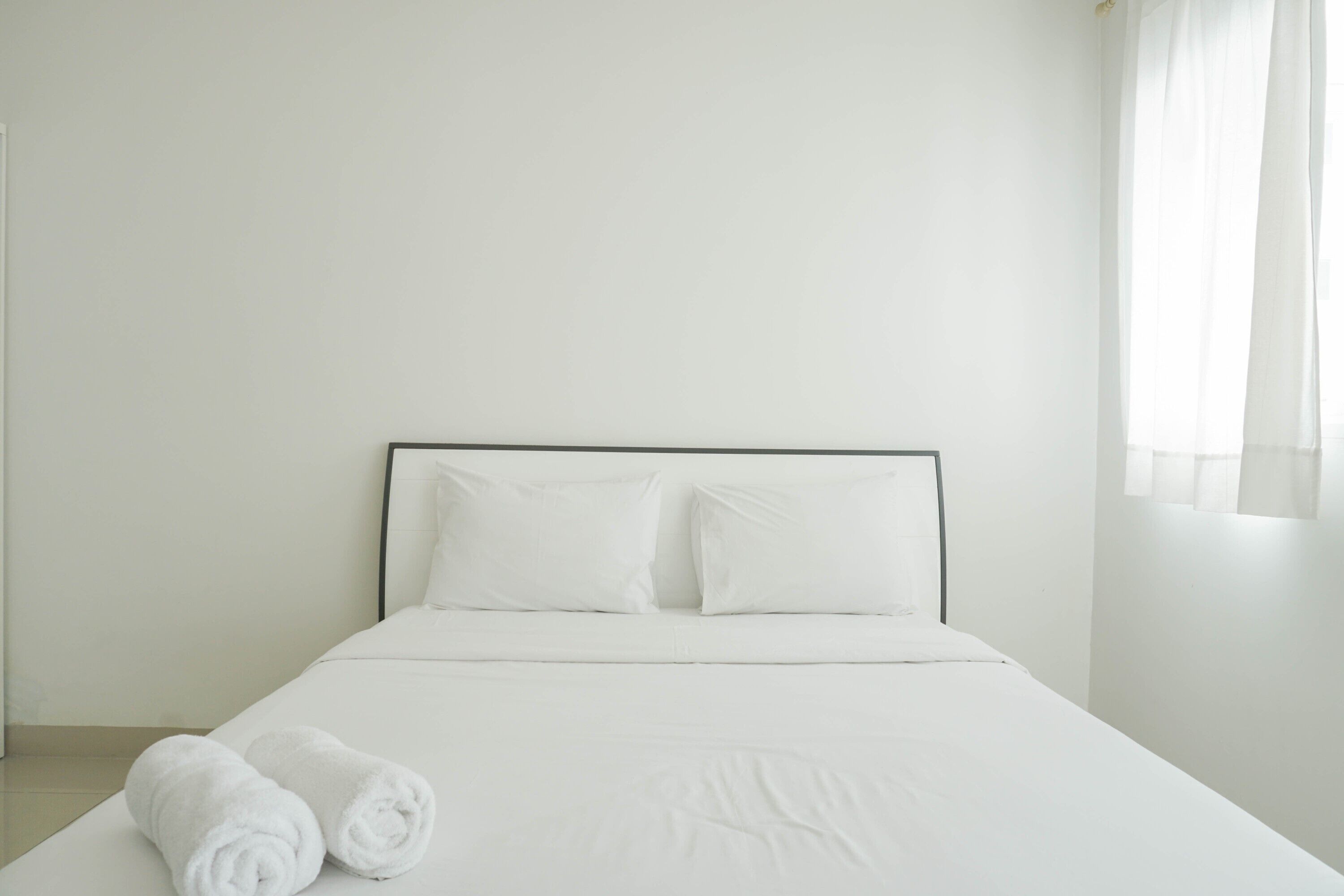 A neatly made bed with white linens and pillows, a rolled white towel on the side, and a plain white wall with a window on the right.