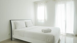 A white, single-bed room with a simple design, featuring a bed with white linens and pillows, a small bedside table, and a window with sheer curtains.