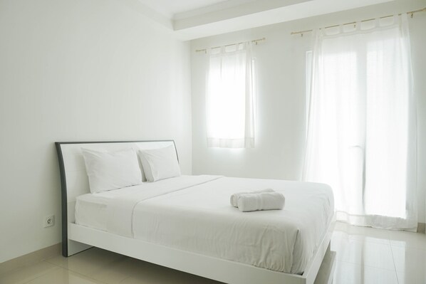 A white, single-bed room with a simple design, featuring a bed with white linens and pillows, a small bedside table, and a window with sheer curtains.