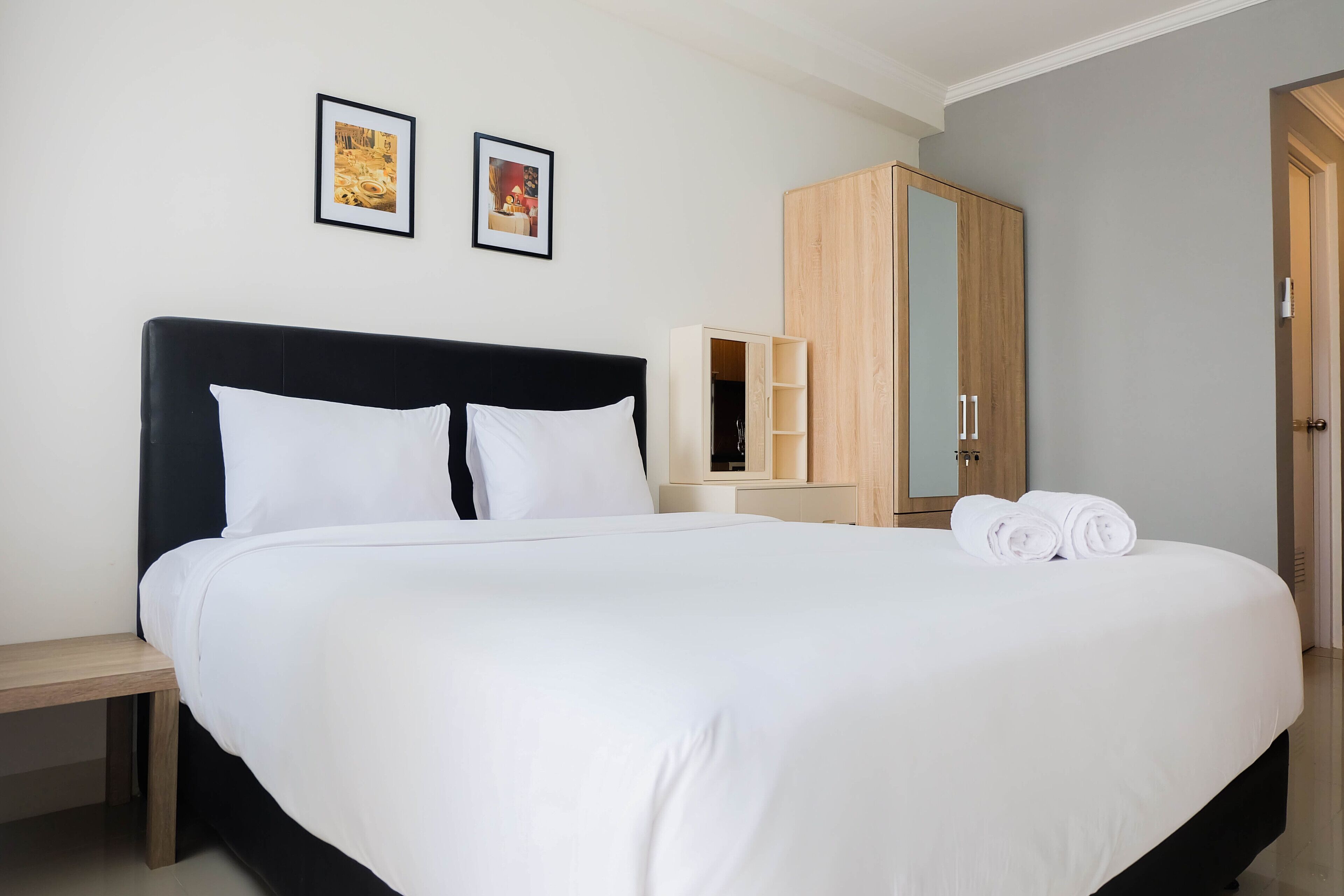 A neatly made bed with white linens, a wooden nightstand, a wardrobe, and two framed pictures on the wall.