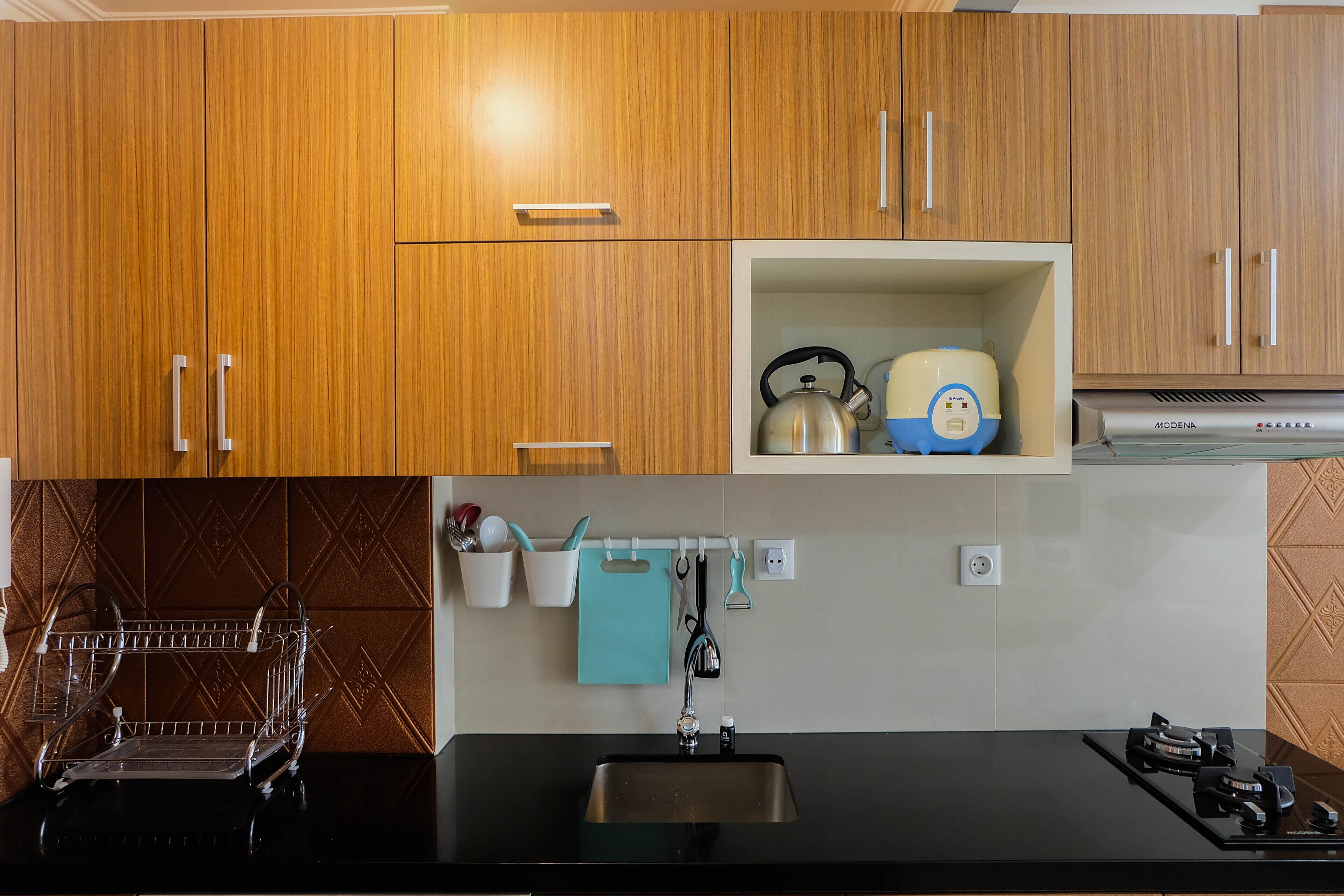 A kitchen with wooden cabinets, a black countertop, a sink, a dish rack, and a small shelf with kitchen utensils.