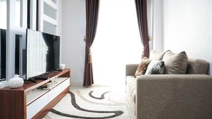 A modern living room with a flat-screen TV, a wooden console table, a beige sofa, and a large window with curtains. - Modern 2Br At Signature Park Grande Apartment (Jakarta)