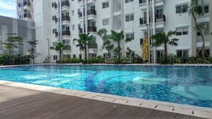 A swimming pool with a wooden deck, surrounded by lush greenery and a multi-story building with balconies. - Comfortable Studio Room At Signature Park Grande Apartment (Jakarta)