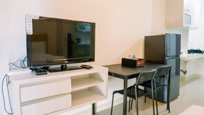 Un espacio habitable compacto con un televisor de pantalla plana sobre un mueble blanco, una pequeña cocina con refrigerador y dos sillas negras junto a una mesa de comedor.