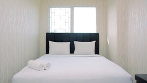 A neatly made bed with white linens and two rolled towels, a dark headboard, and a window with a geometric pattern. - Brand New And Modern 1Br Signature Park Grande Apartment (Jakarta)